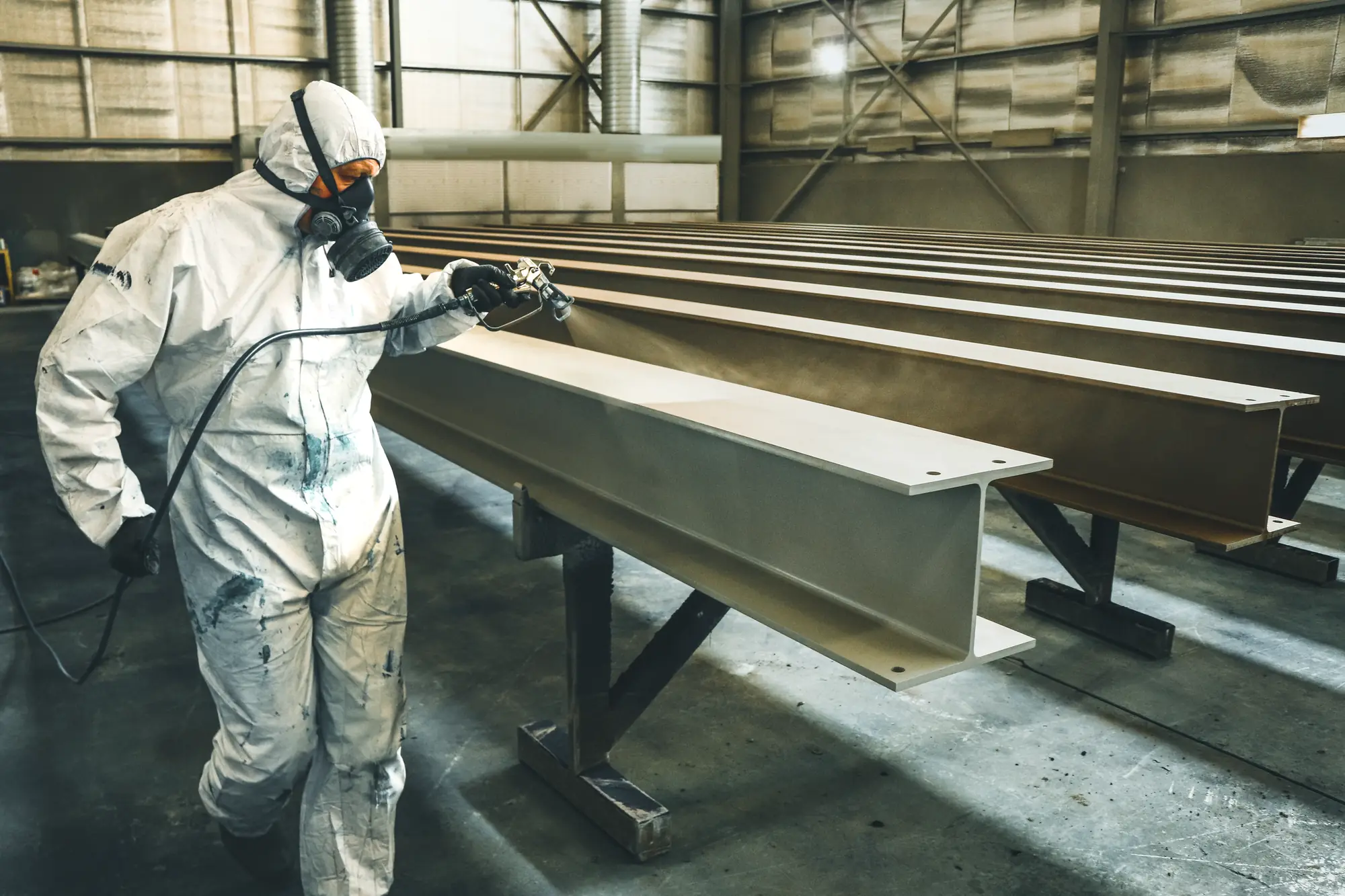 Worker in a white disposable coverall and half-face respirator spray-coating structural steel I-beams in a fabrication shed