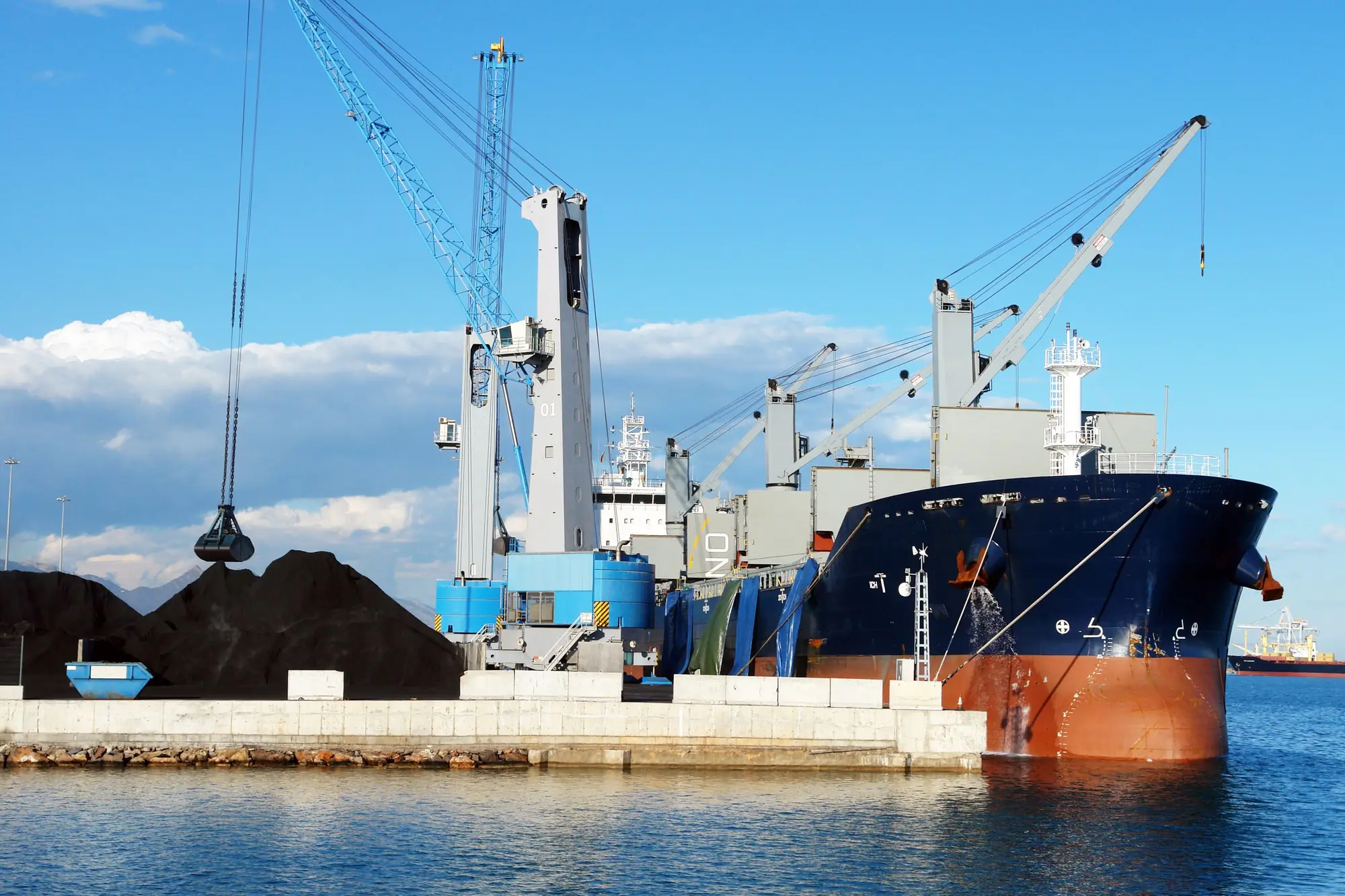 Bulk carrier moored at a quay beside dark mineral stockpiles, with a crane grab and the ship's deck cranes under a blue sky