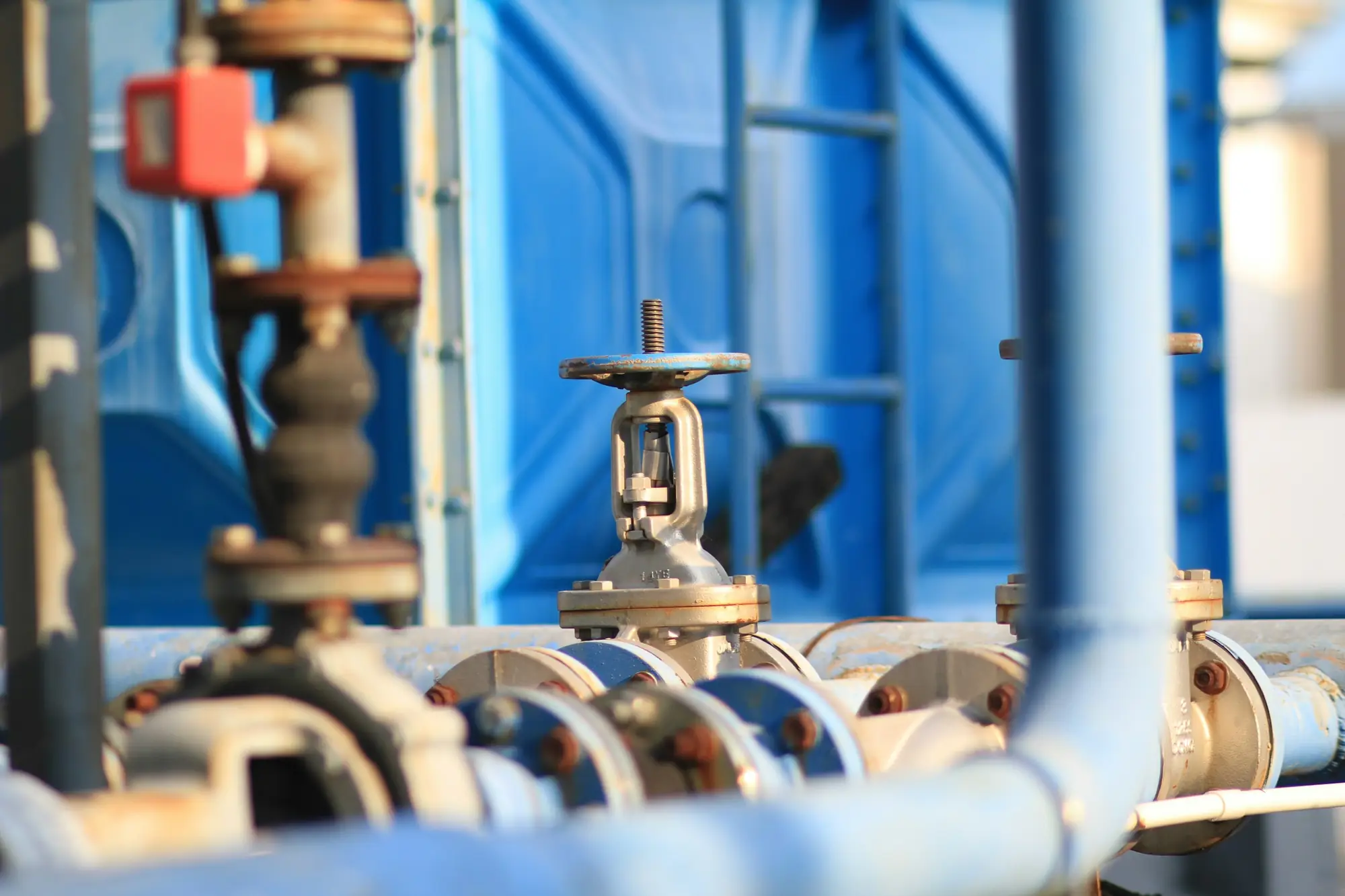 Flanged gate valve on light-blue water pipework in front of a blue sectional storage tank